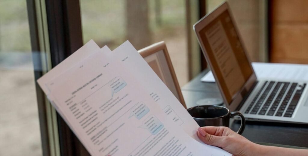 Person holding and reviewing documents near window with laptops on a desk.